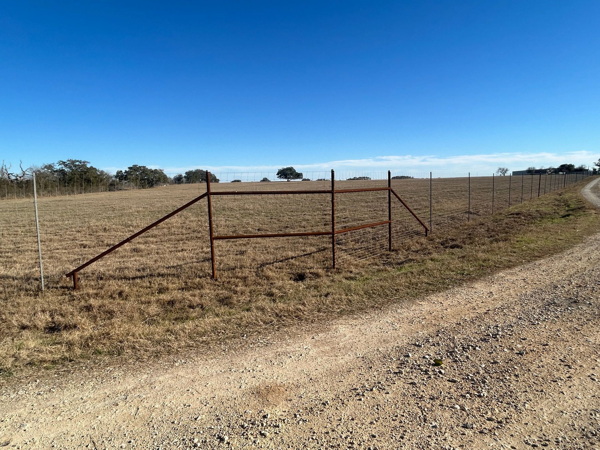 Rusty metal fence along a dry field beside a gravel road under a clear blue sky