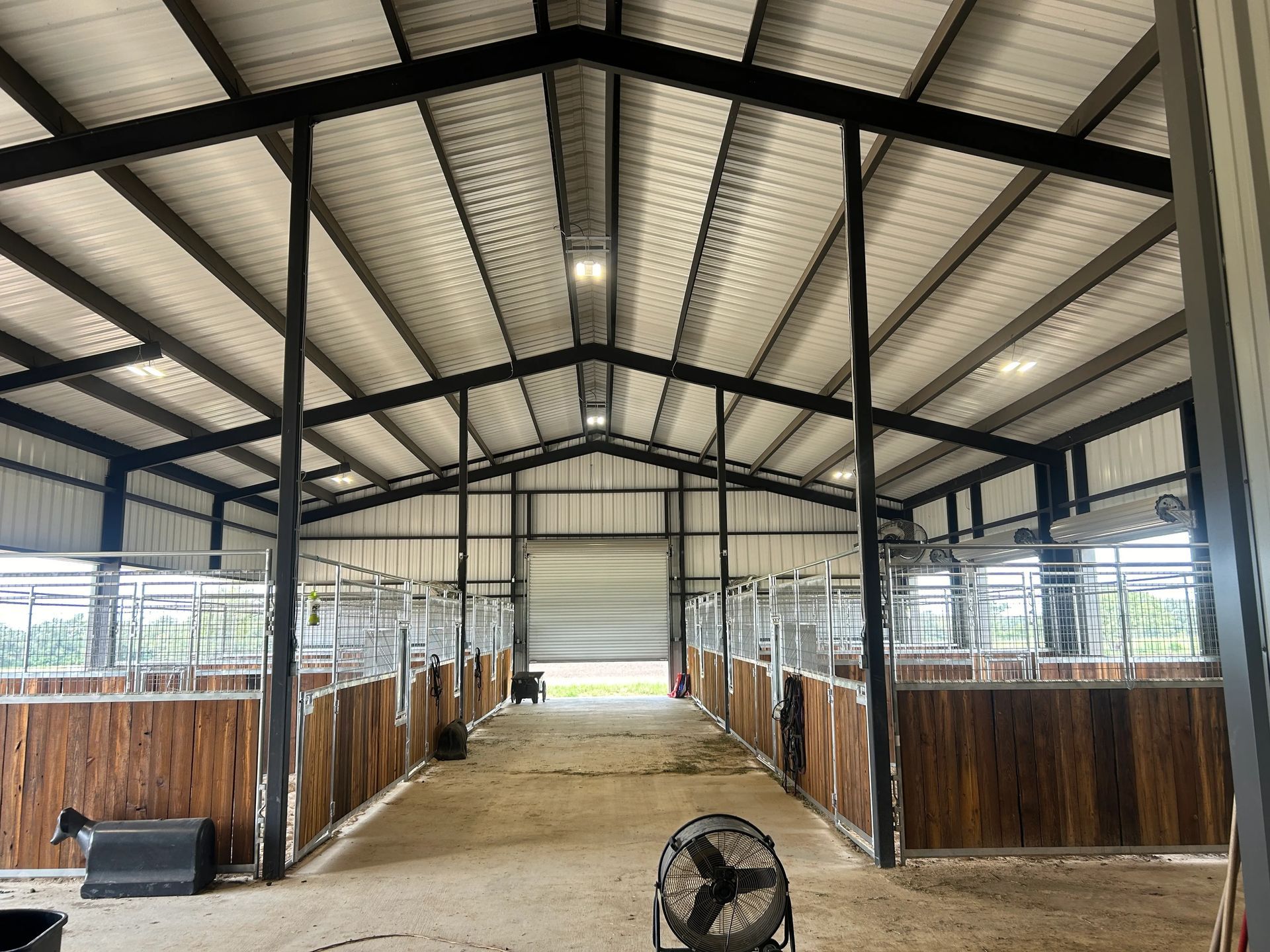 Covered livestock barn with metal roof, wood stalls, and a central dirt aisle, viewed from the entrance