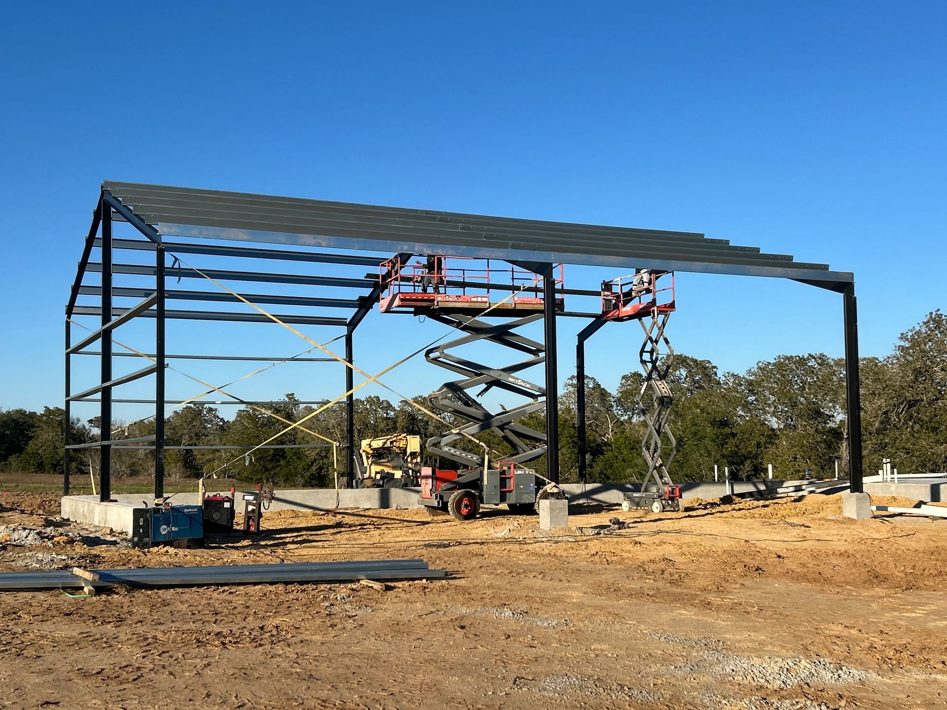 Steel building frame under construction on a dirt lot, with scaffolding and a red lift beneath a blue sky