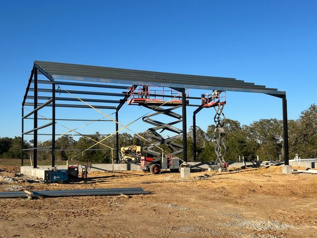 Steel building frame under construction on a dirt lot, with scaffolding and a red lift beneath a blue sky