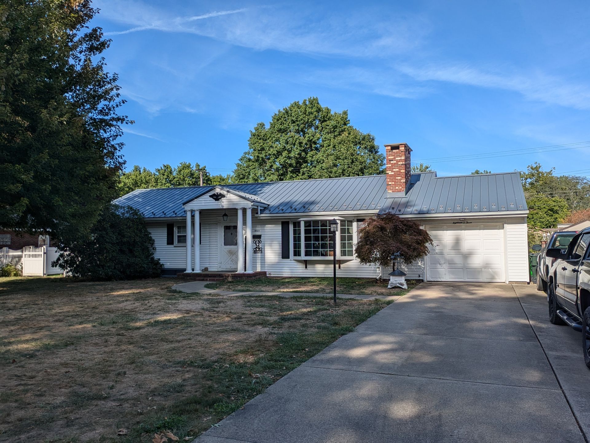 Single-story white house with a blue metal roof, front porch, driveway, and parked car under a blue sky