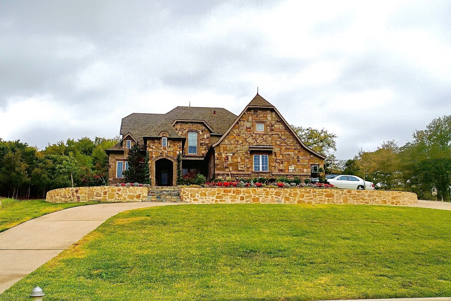 Stone house with a curved driveway, grassy lawn, and low stone wall under a cloudy sky. A white car is parked in front.