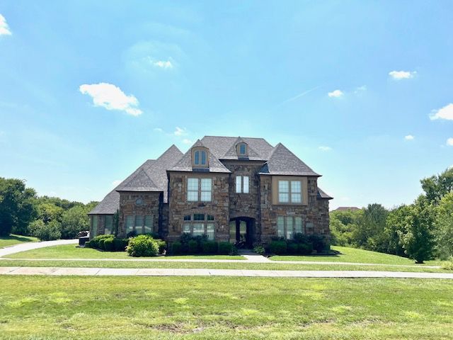 Two-story stone house with multiple gables, windows, and a dark roof on a grassy lawn under a blue sky.