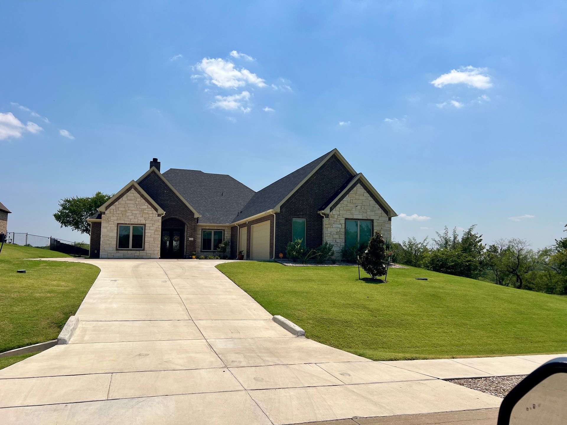 Tan stone house with a dark roof and a concrete driveway on a sunny day with a blue sky.
