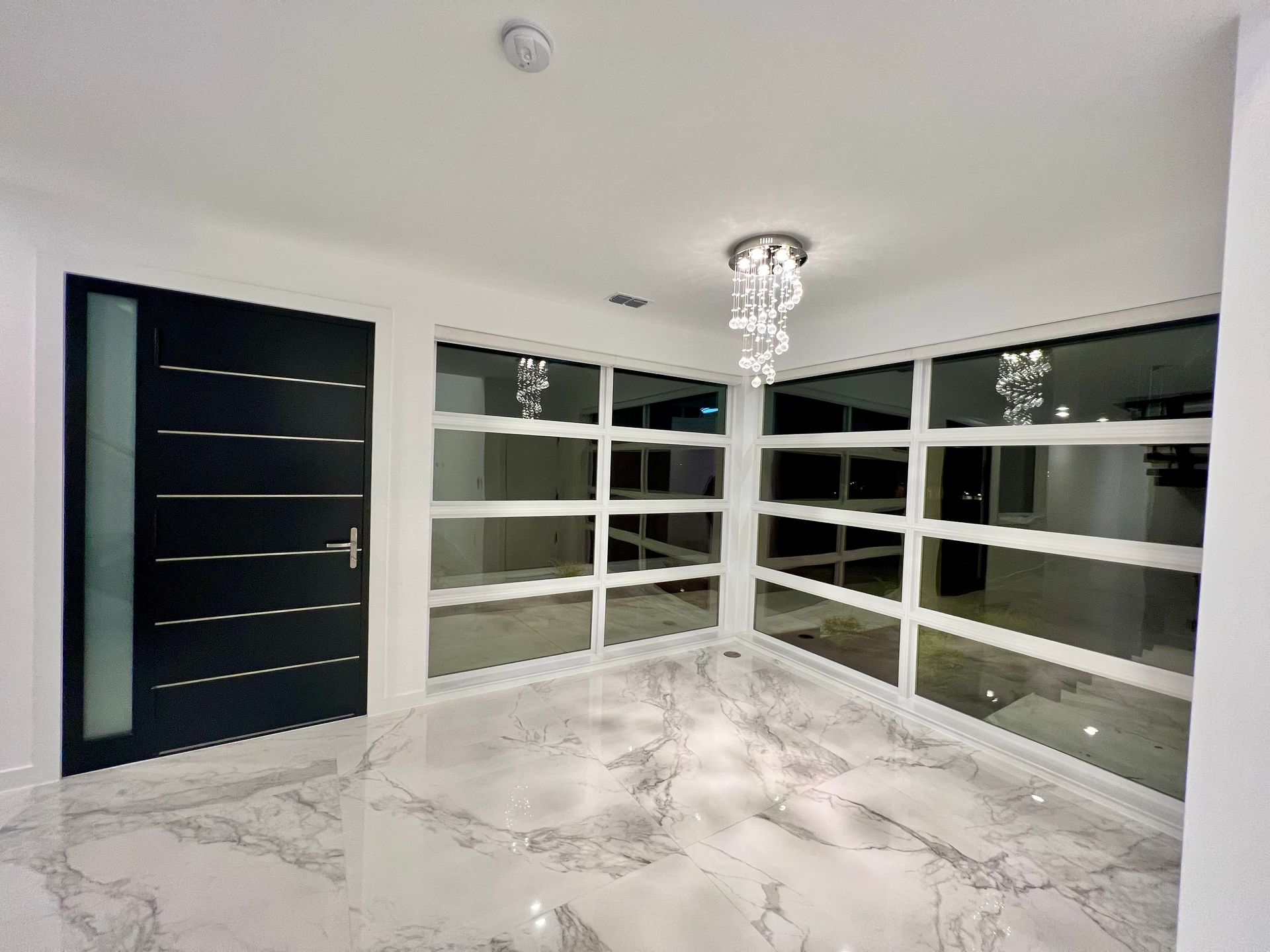 A modern entryway with a black door, marble flooring, and large windows, showcasing a spiral chandelier.