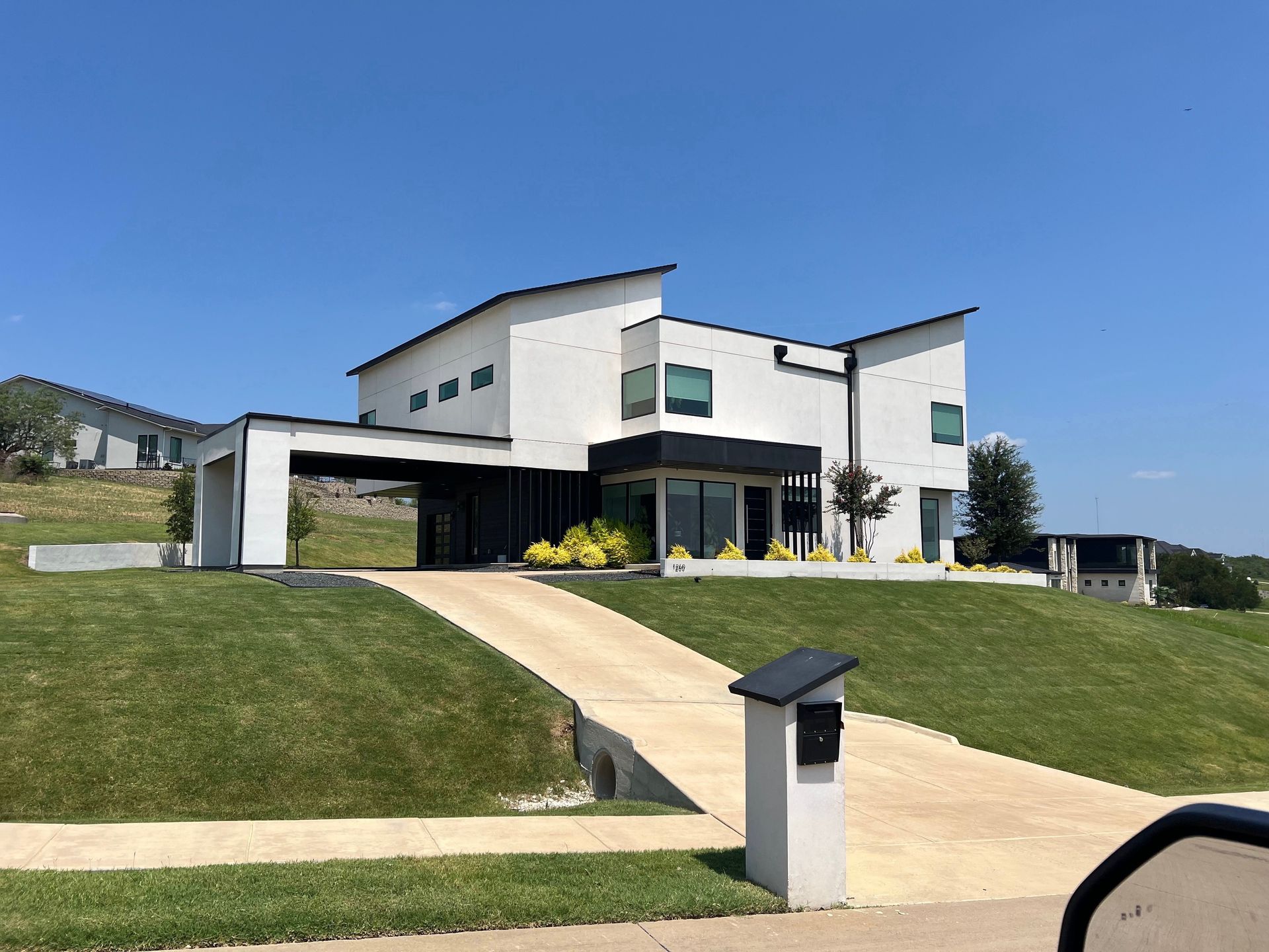 Modern white house with black trim, a concrete driveway, and green lawn under a blue sky. A mailbox is in the foreground.
