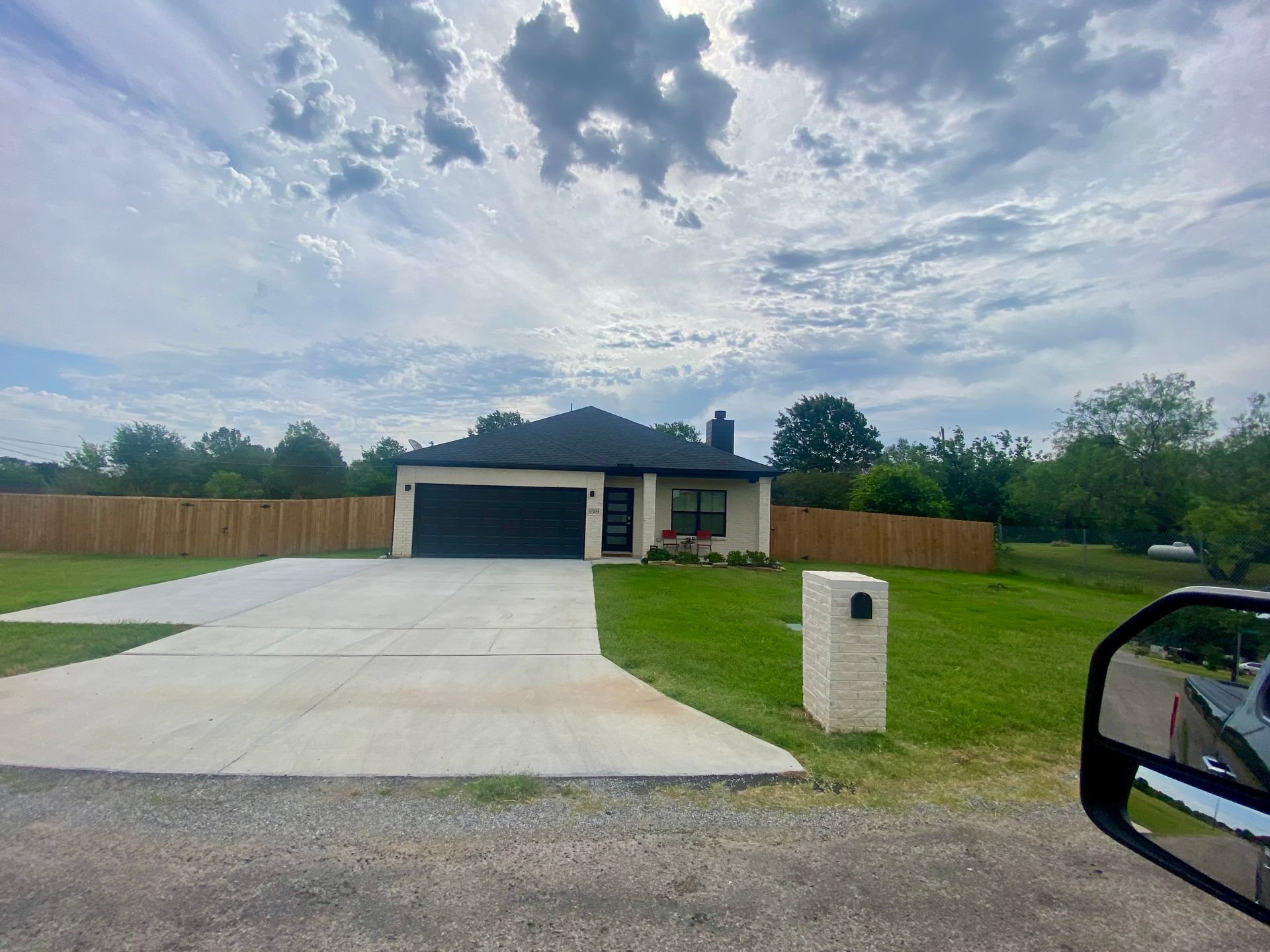 A modern house with a dark garage door and a concrete driveway, set against a cloudy sky and wooden fence.