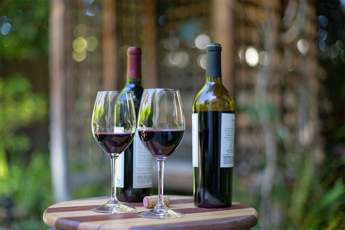 Two wine glasses with red wine and two wine bottles on a wooden table, outdoor setting.