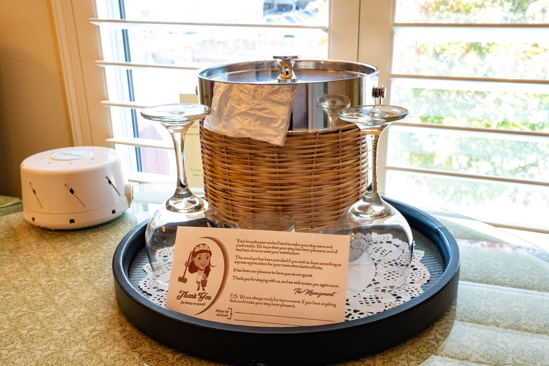 Wine glasses and ice bucket on a tray with a note, near a window and white decor.