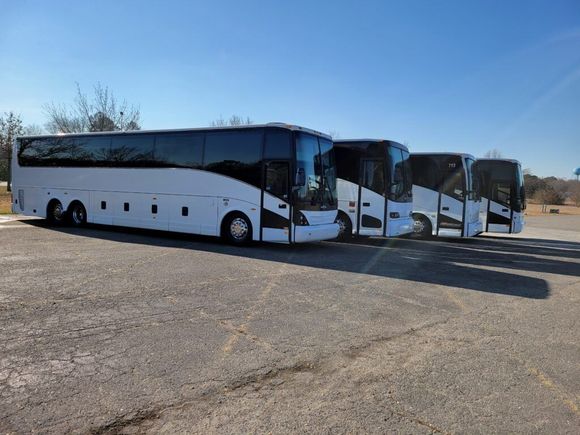 Four white passenger buses parked in a lot under a clear blue sky.