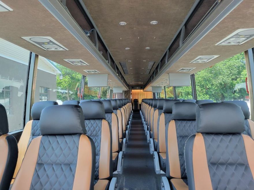 Interior of a bus with rows of black and tan seats, overhead compartments, and windows.