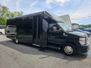 Black party bus parked on asphalt, door open. Blue sky, other vehicles and building visible.