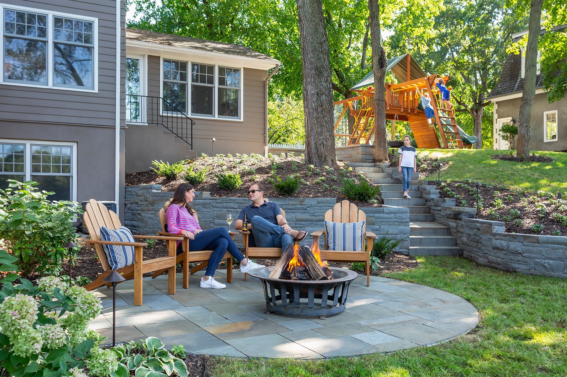 A young couple sitting at a round patio with firepit while children enjoy playset in background
