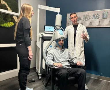Clinic exam with seated person wearing a head-mounted device, while two staff members stand nearby.