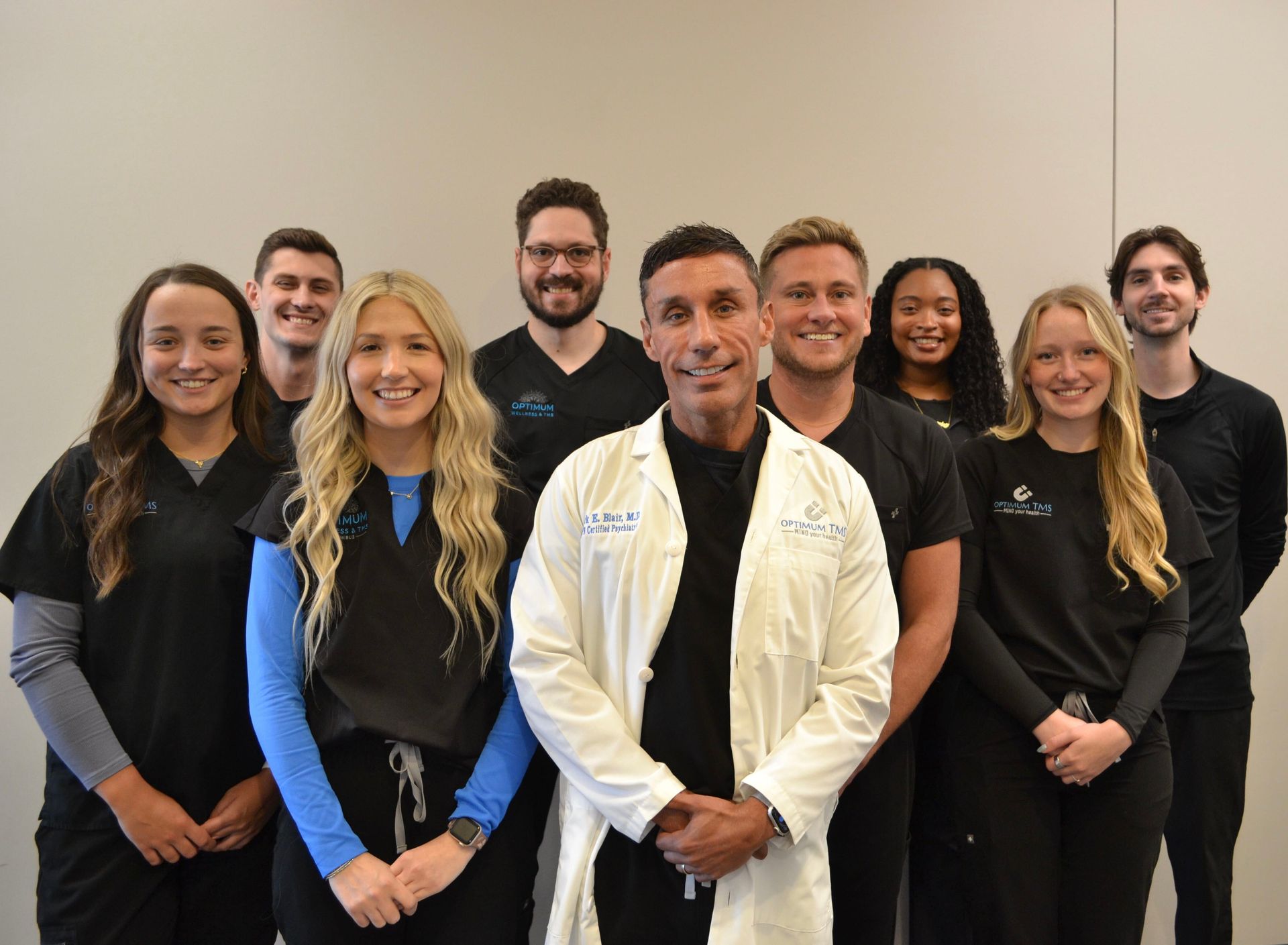 Eight people posing indoors, smiling, in black outfits with one man in a white jacket front and center