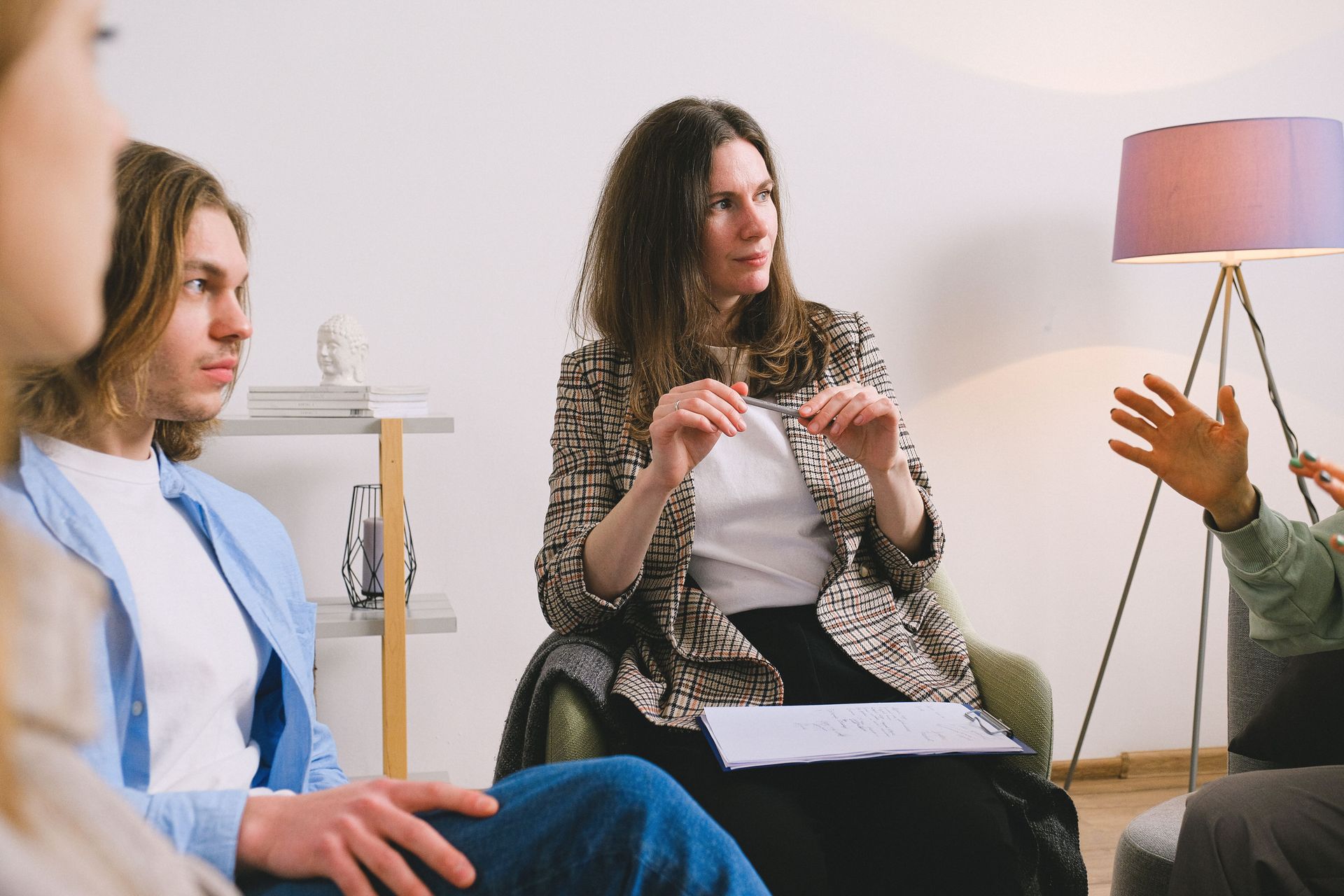 Three people in a bright room, one woman speaking while holding papers, two others listening.