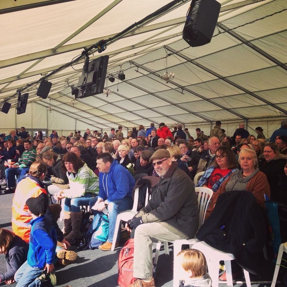 Large crowd seated under a tent, listening. Speakers mounted on the ceiling.