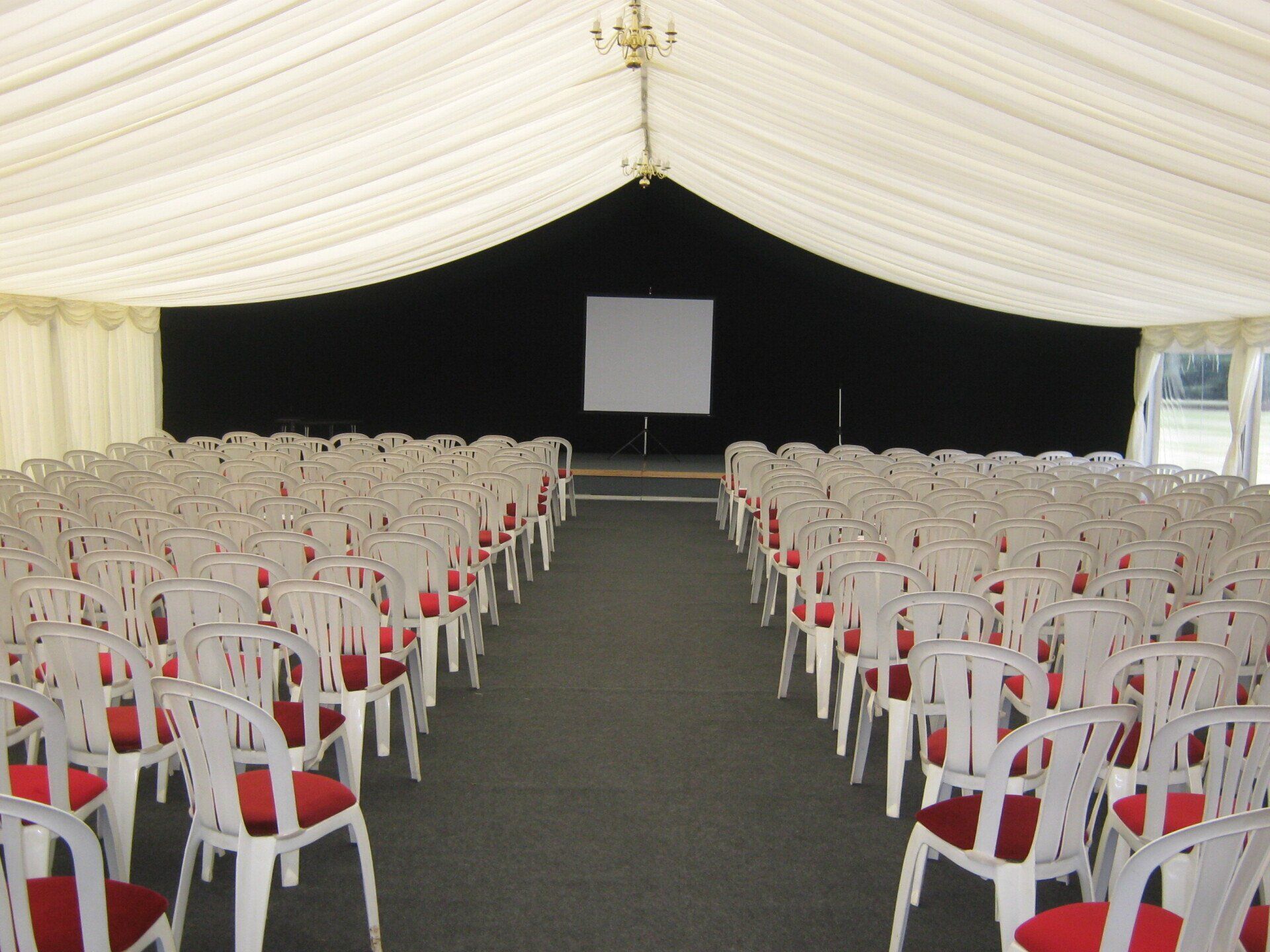 Rows of white chairs with red cushions face a stage with a screen inside a large tent.
