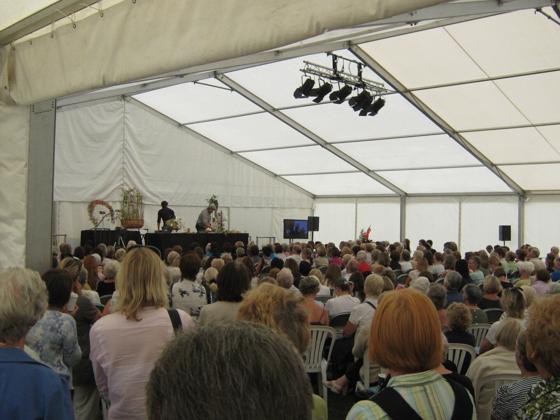 Large crowd attending an event inside a white tent. Speakers on a stage; stage lights are overhead.