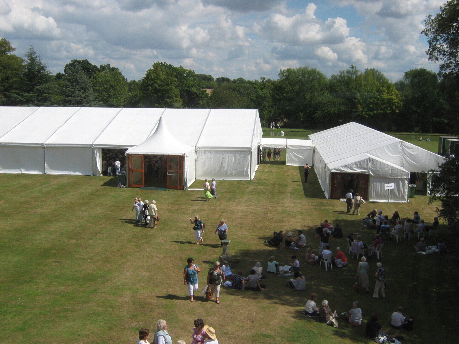 Large white tents set up on a grassy field with people milling about, trees in the background, under a cloudy sky.