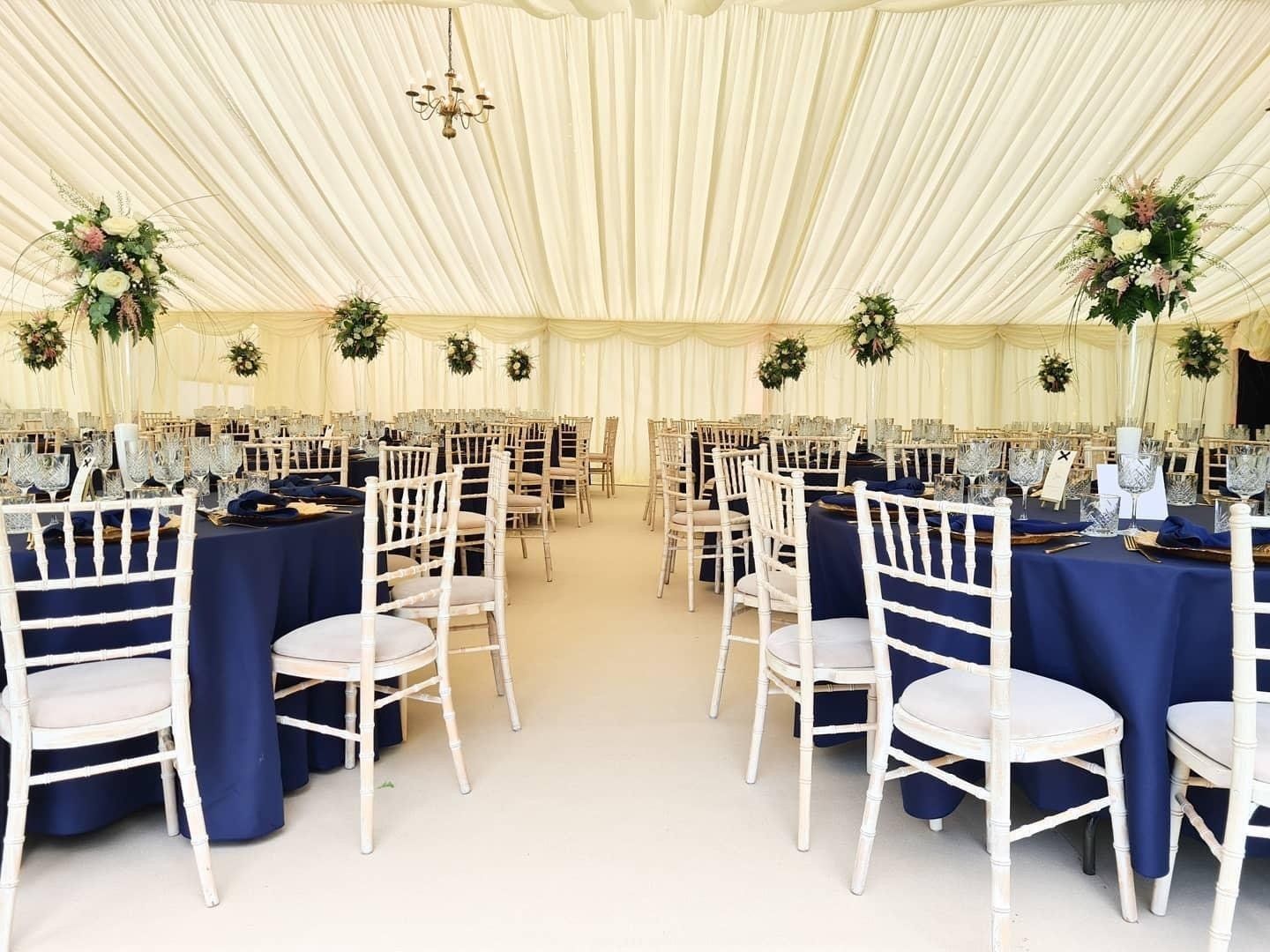 Wedding reception tent with navy tablecloths, white chairs, and floral centerpieces.
