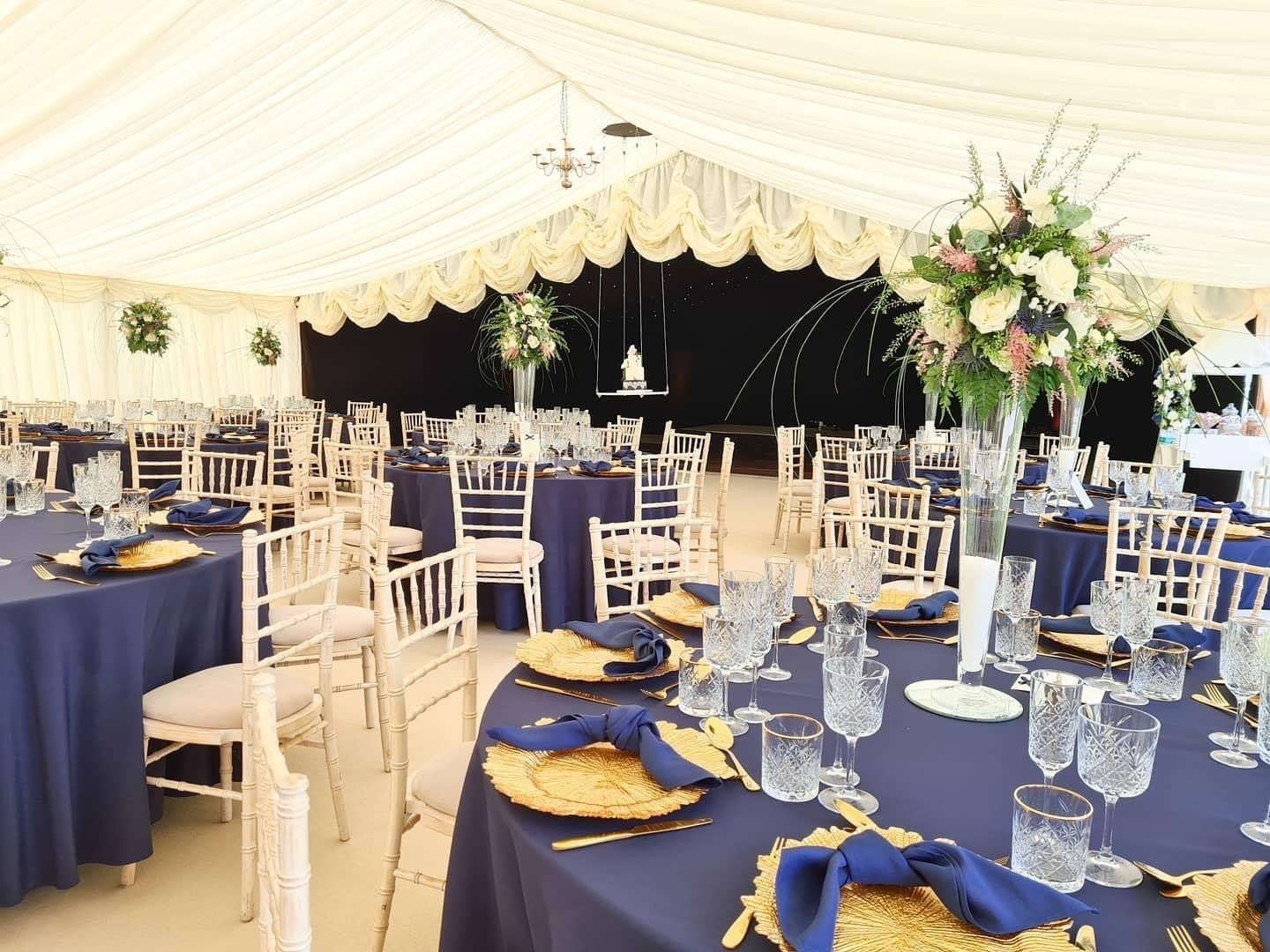 Wedding reception: tables set with blue tablecloths, gold chargers, floral centerpieces under a tent.