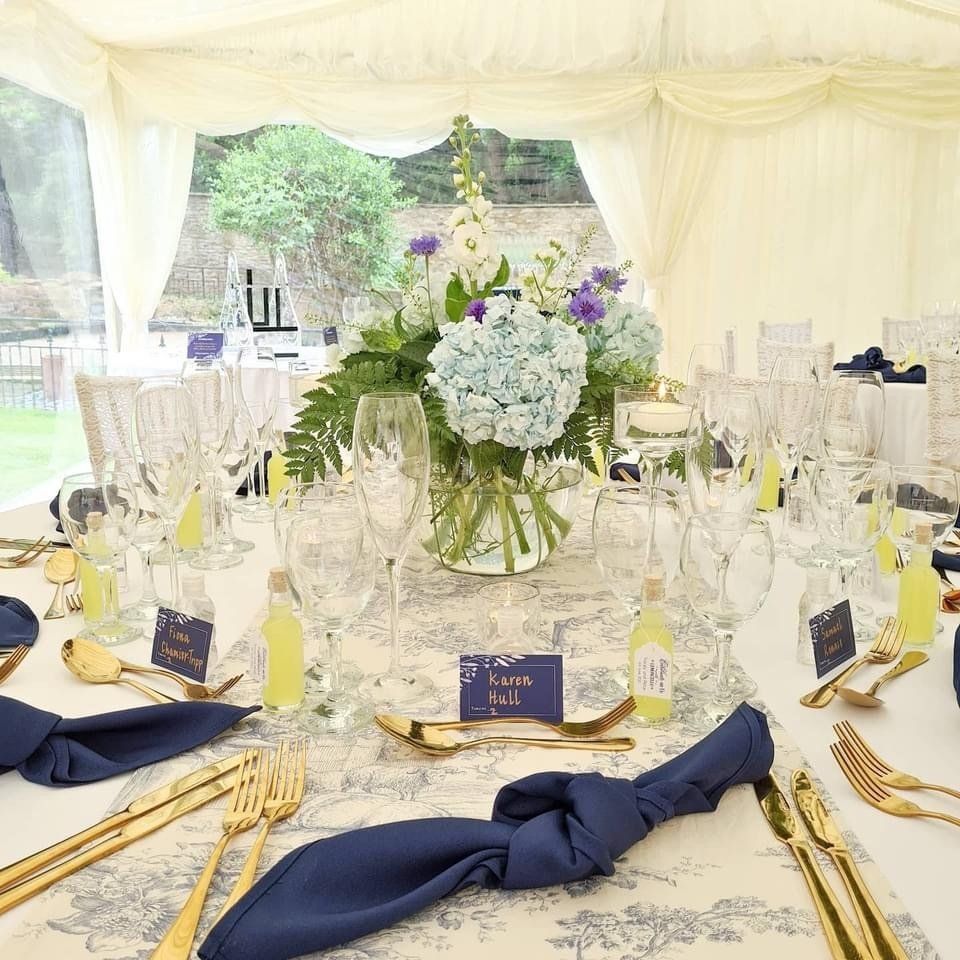 Wedding reception table set with gold cutlery, navy napkins, and floral centerpiece.