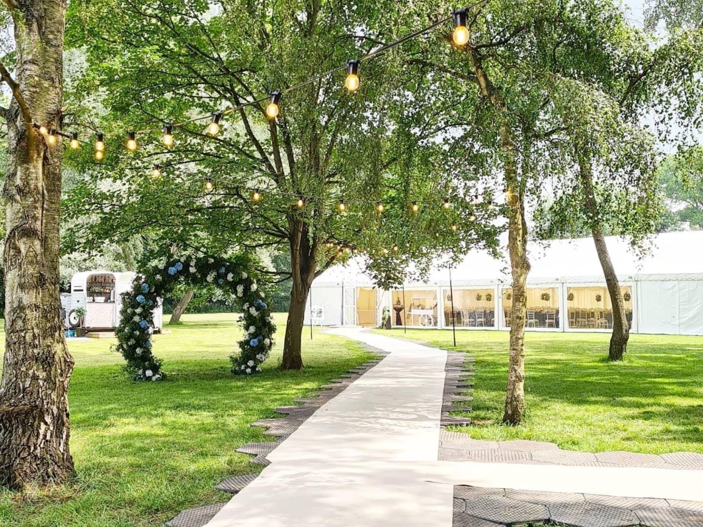 Pathway leading to a white tent in a green space, with a floral arch and string lights.