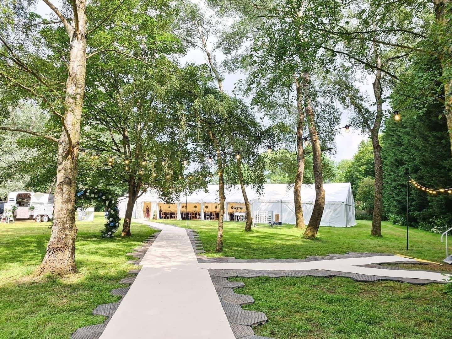 Pathway through a park, leading to a white tent. Trees line the path, green grass surrounding.