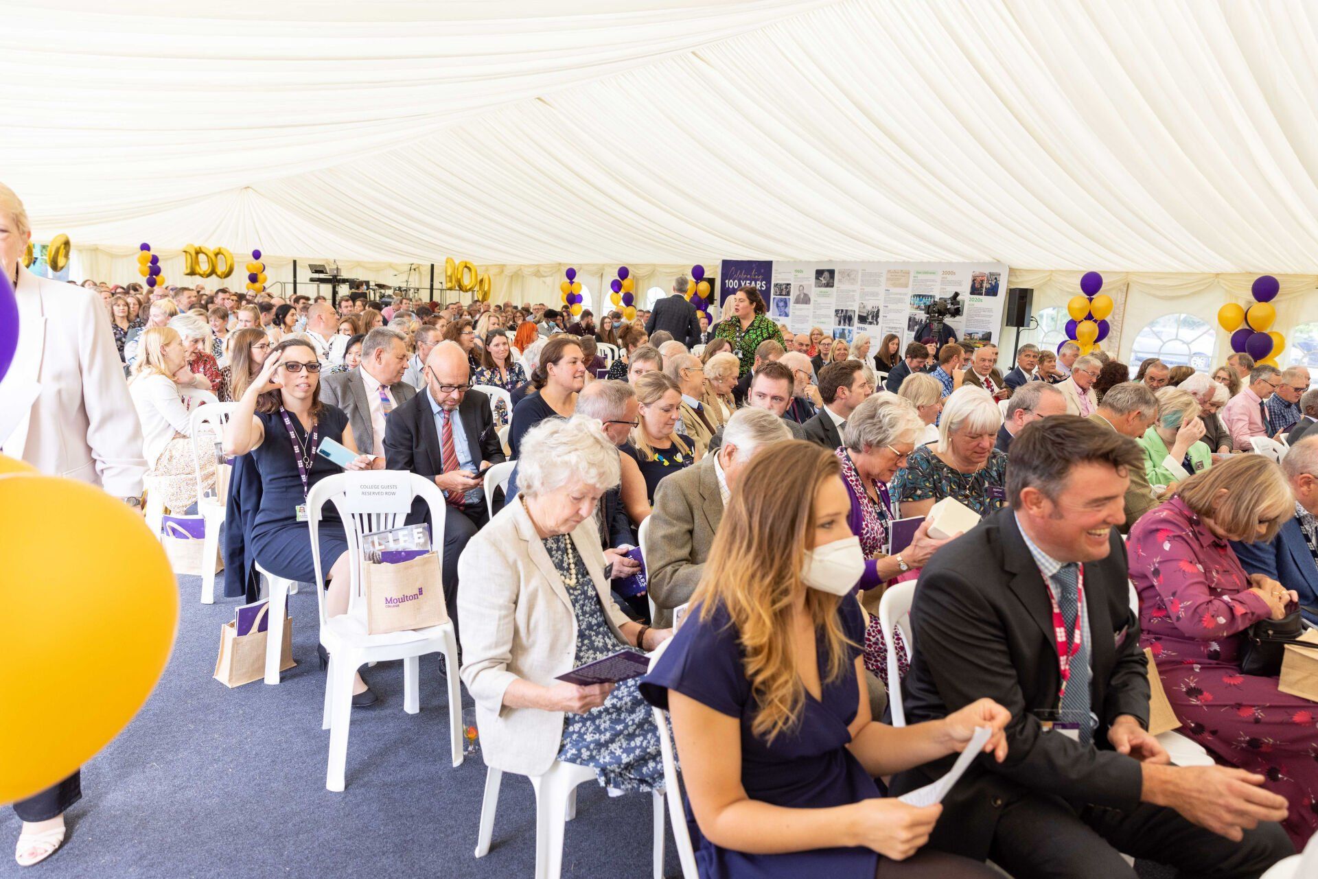 People seated in a tent at an event; yellow and purple balloons, formal attire, white chairs, and ceiling.