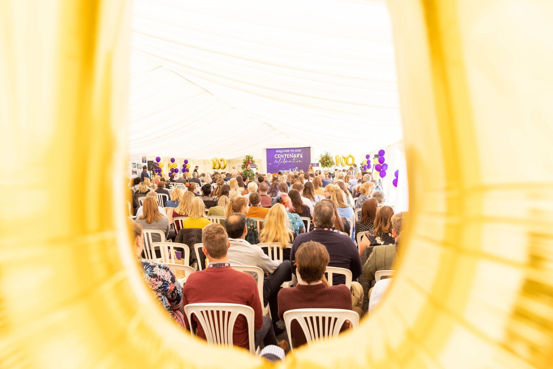 View of an audience sitting in white chairs at a large event; framed by a yellow archway.
