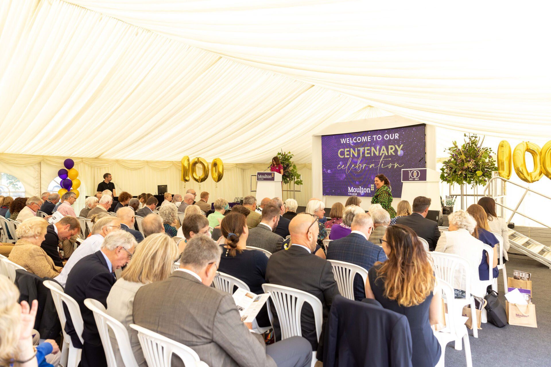 Audience seated at an event inside a tent. A speaker stands before a banner celebrating a centenary.