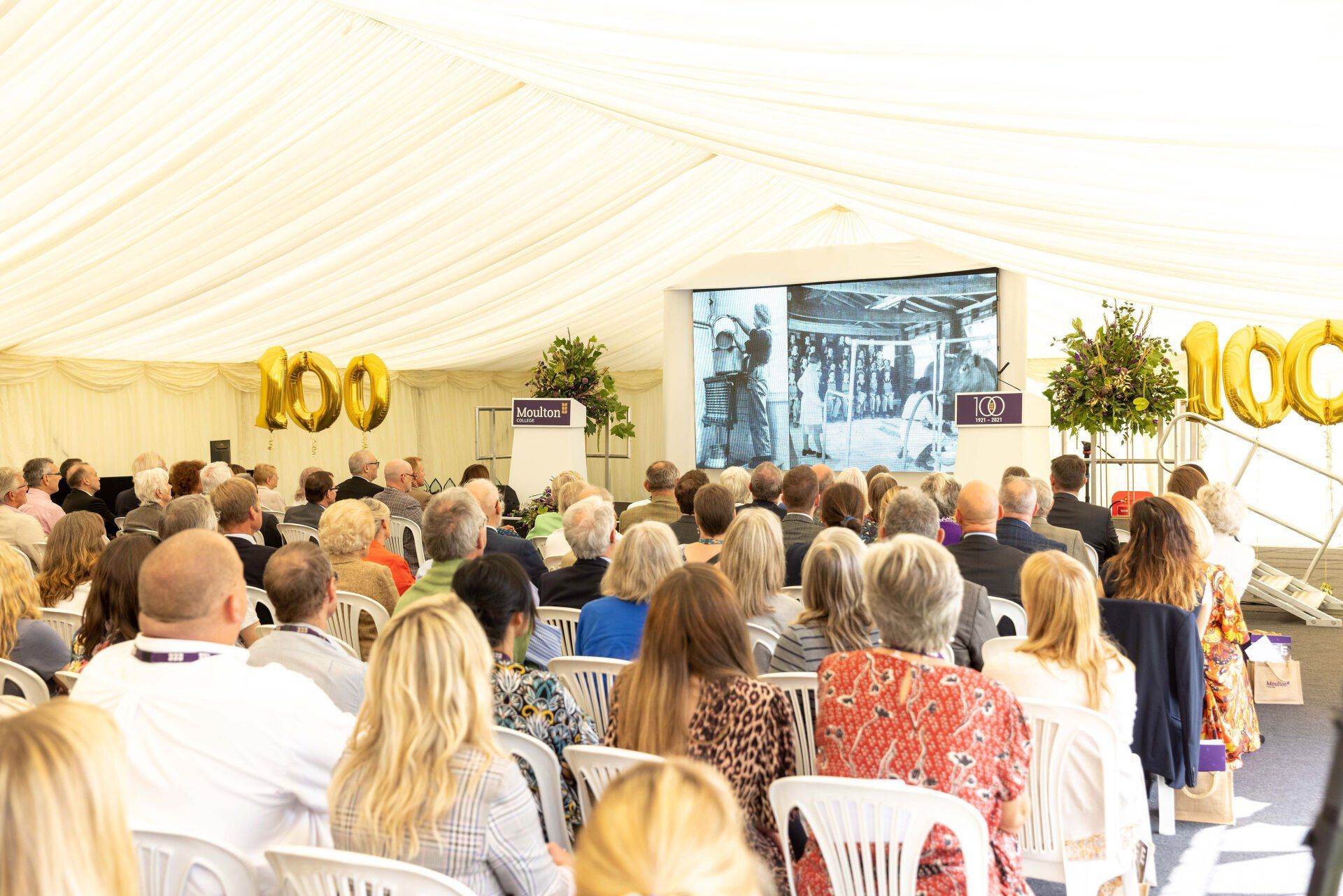 Audience in a tent watching a presentation on a large screen. Gold