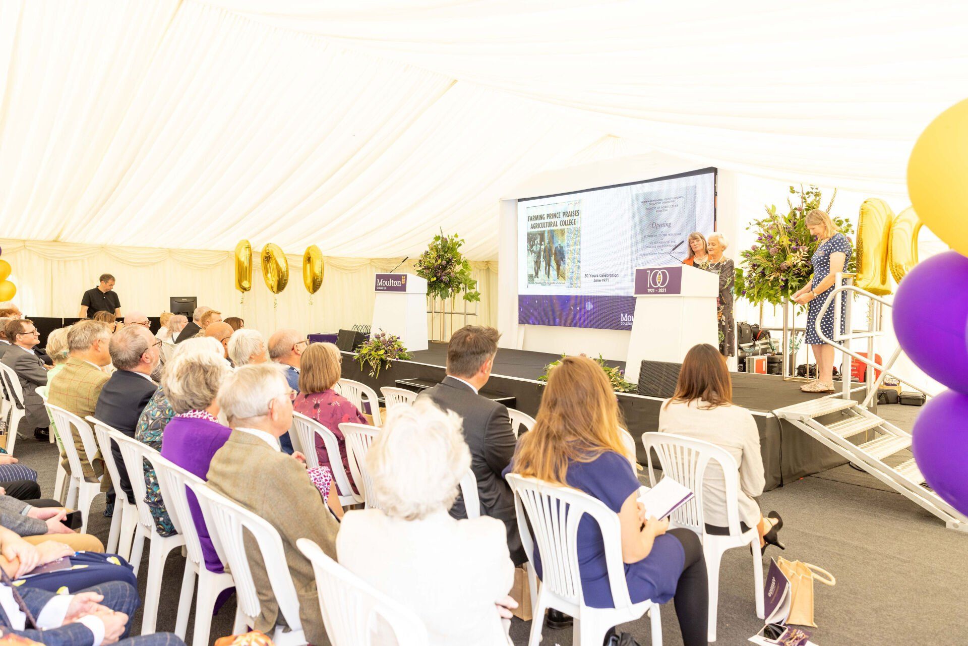 People seated in a tent, listening to a presentation on stage. Gold and purple balloons are visible.