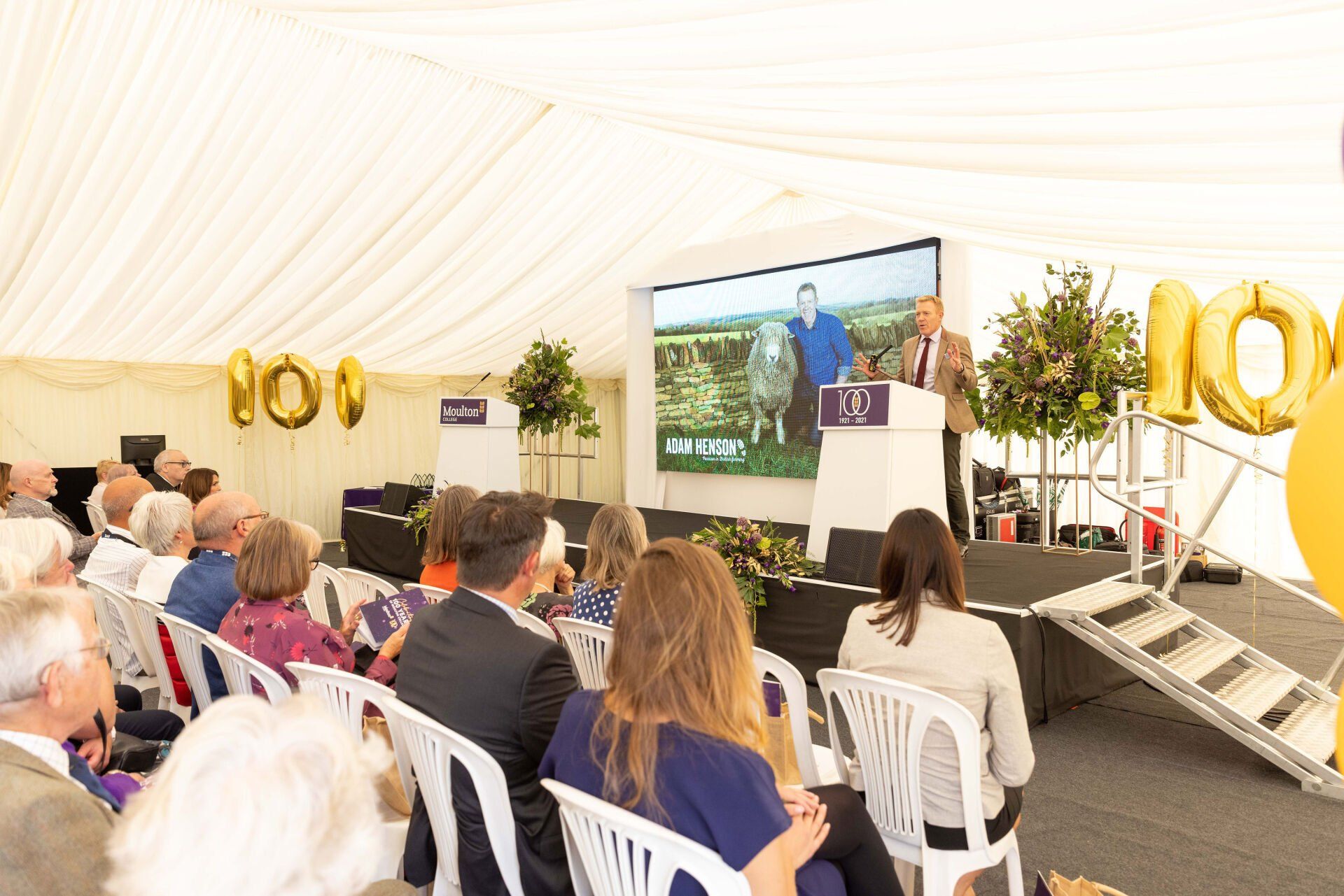 Man speaking at a podium in a tent, large screen behind showing another man. Audience seated.