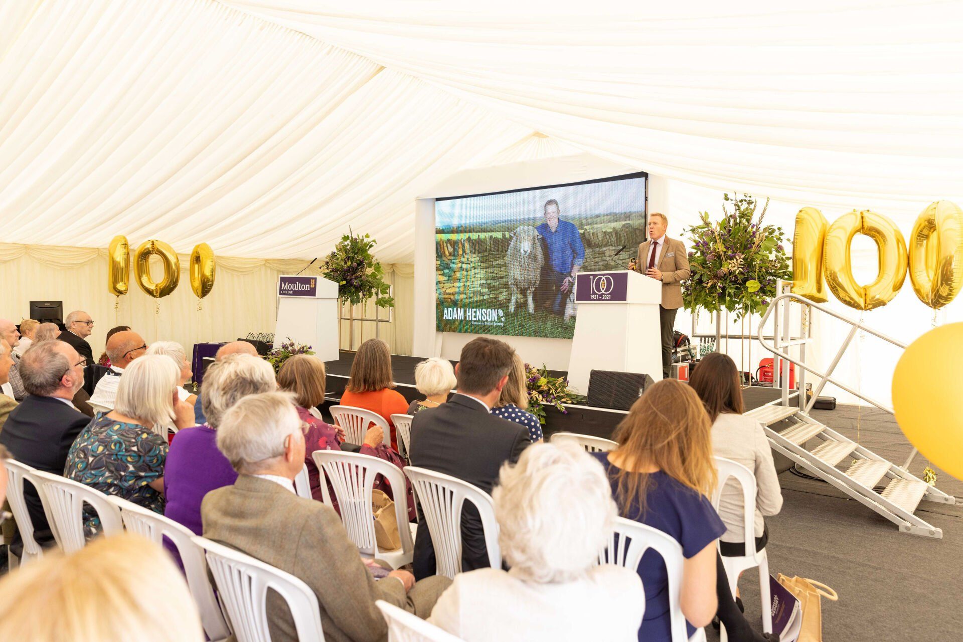 People seated, listening to a speaker at an event under a tent, with a screen and