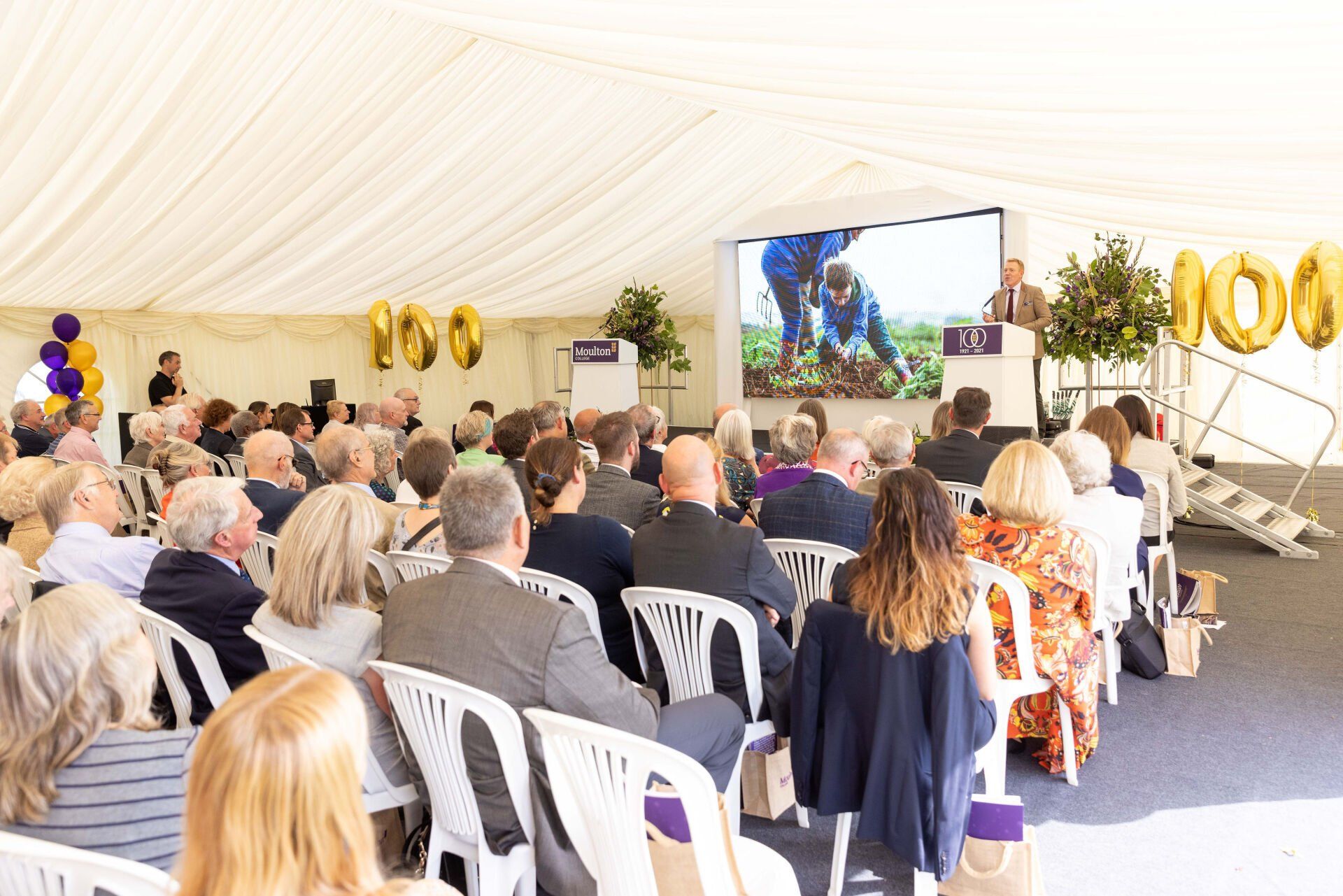 Audience in tent watches speaker at podium with screen displaying people working, celebrating an event.