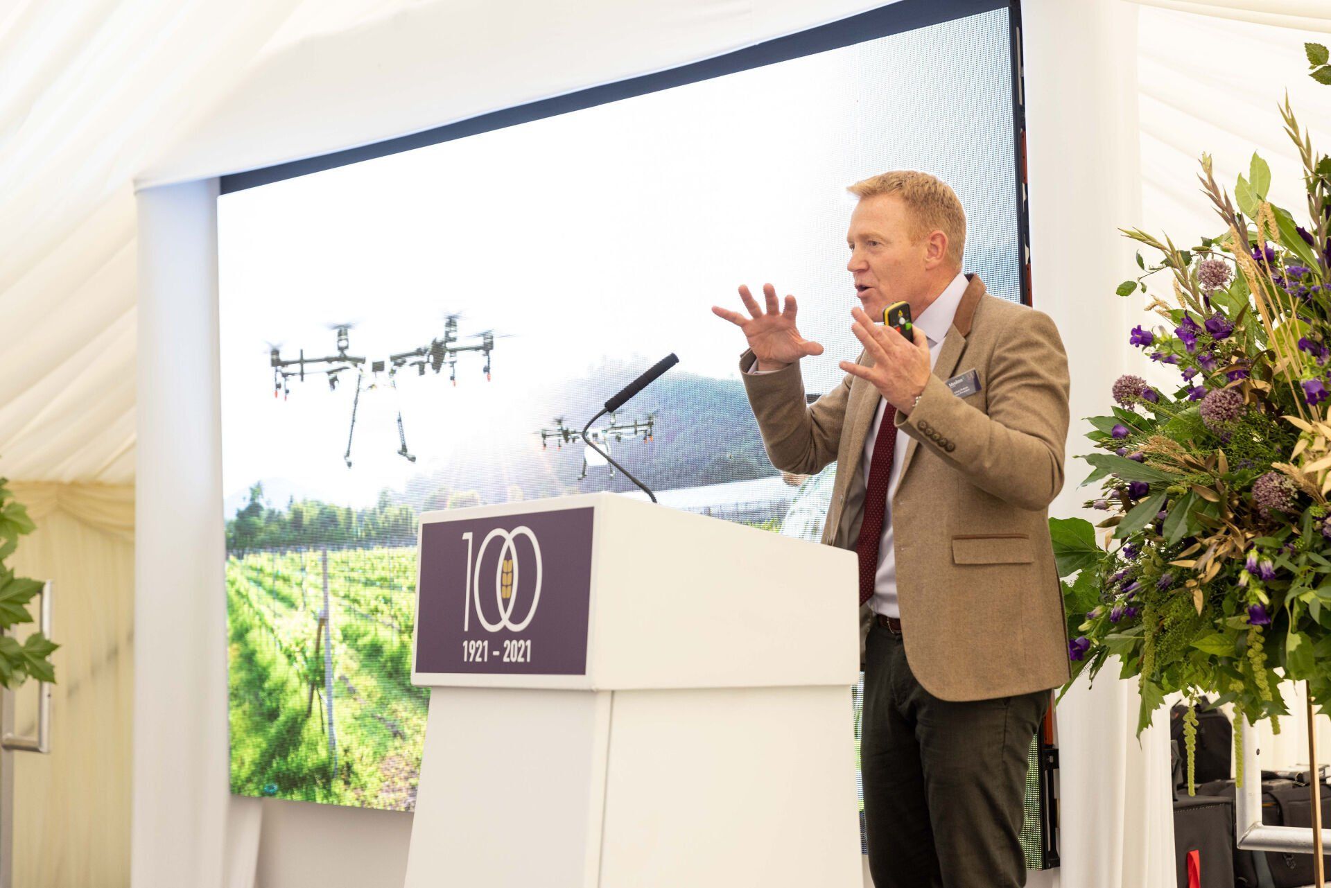 Man speaking at podium, gesturing at screen with drone image. Event in tent, purple flowers on the right.