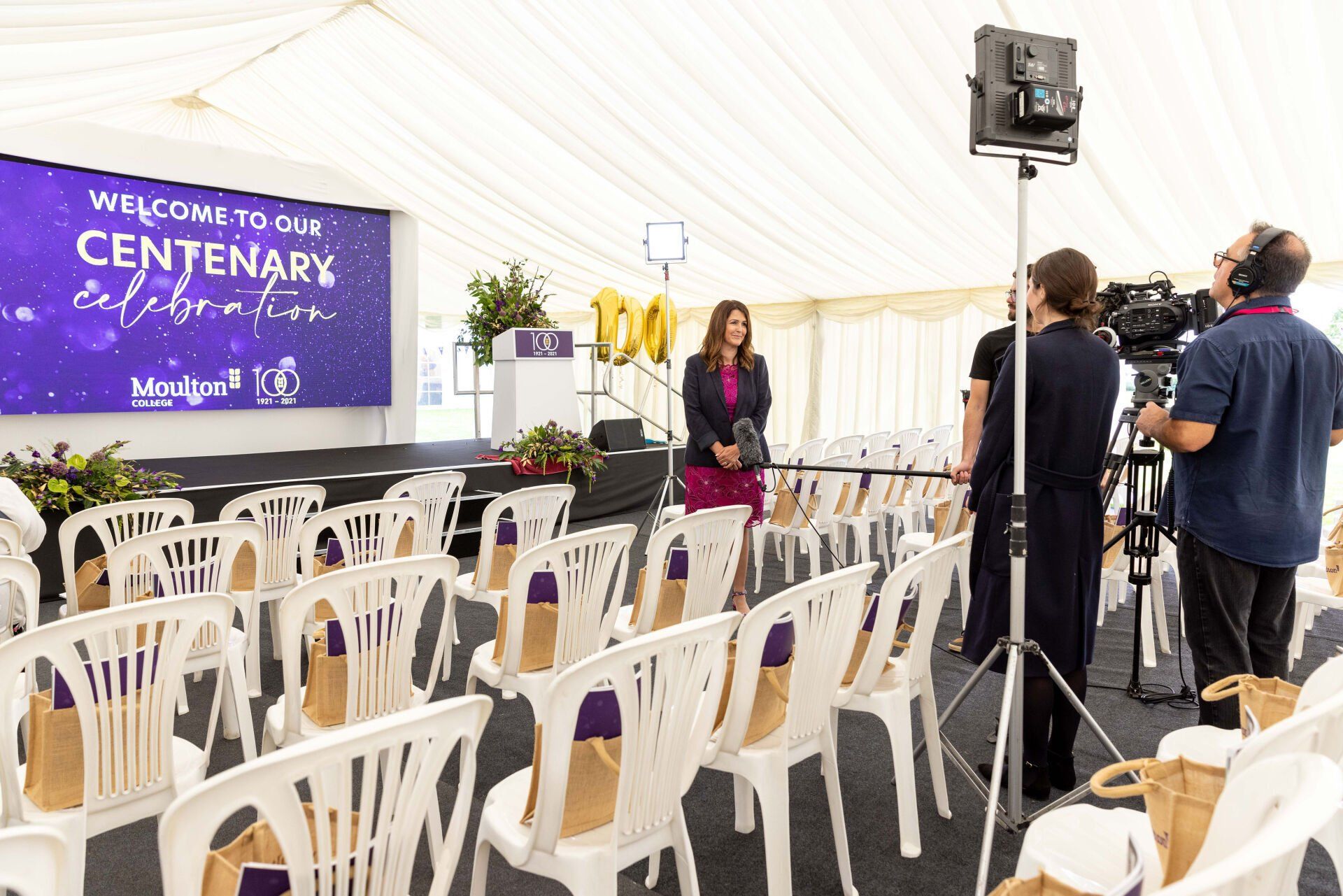 Inside a tent, a woman in a magenta outfit stands on a stage. Crew members film, chairs face the stage.