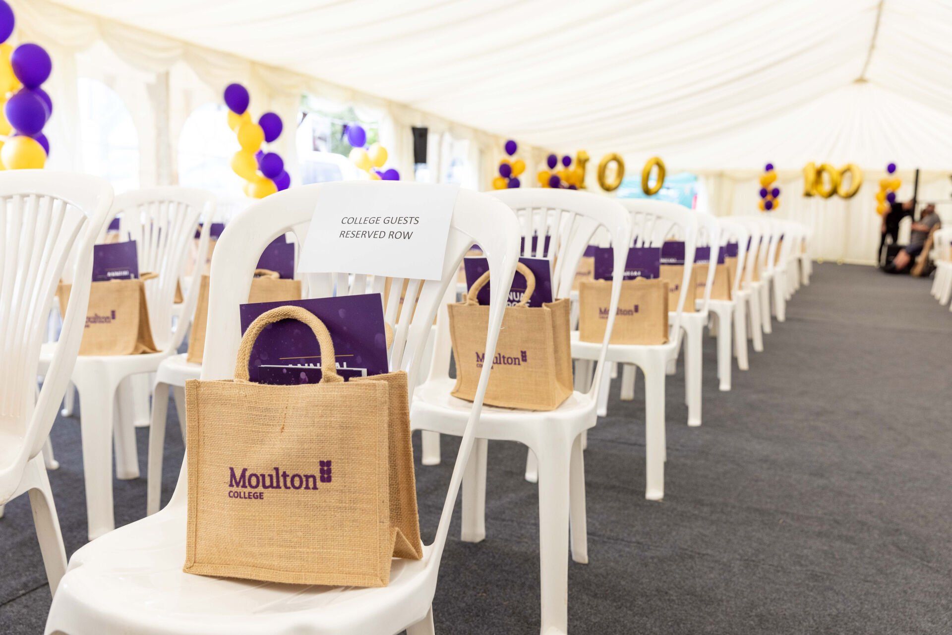 Rows of chairs with gift bags, set up in a tent decorated with purple and yellow balloons.