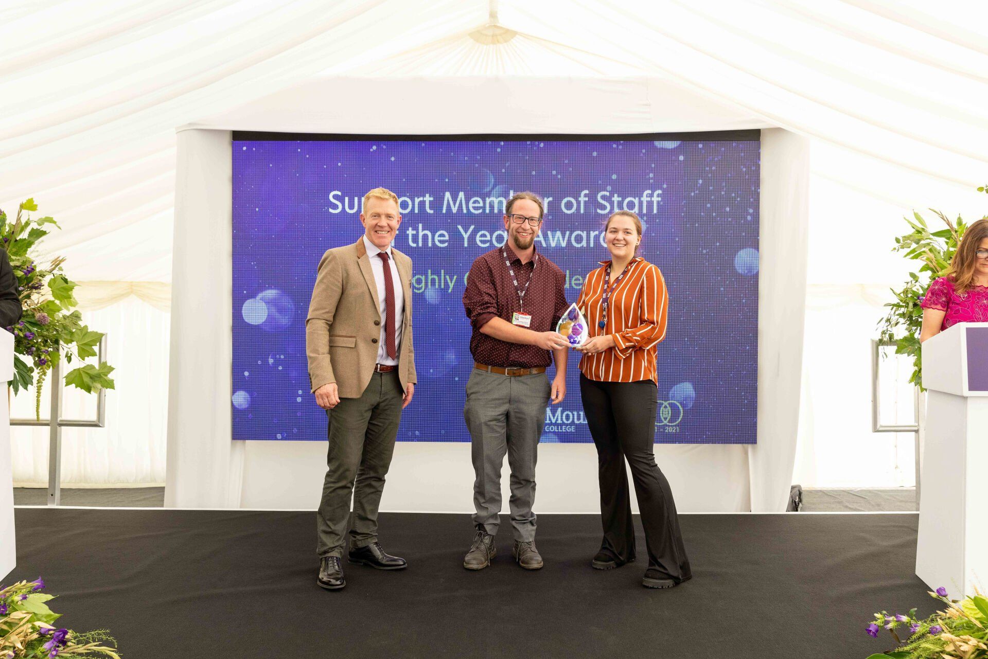 Man receiving an award on stage; two people stand beside him in a white tent.