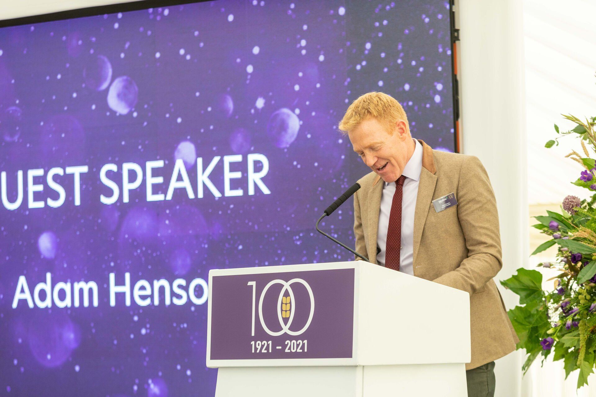 Adam Henson speaking at a podium with a purple backdrop, smiling.