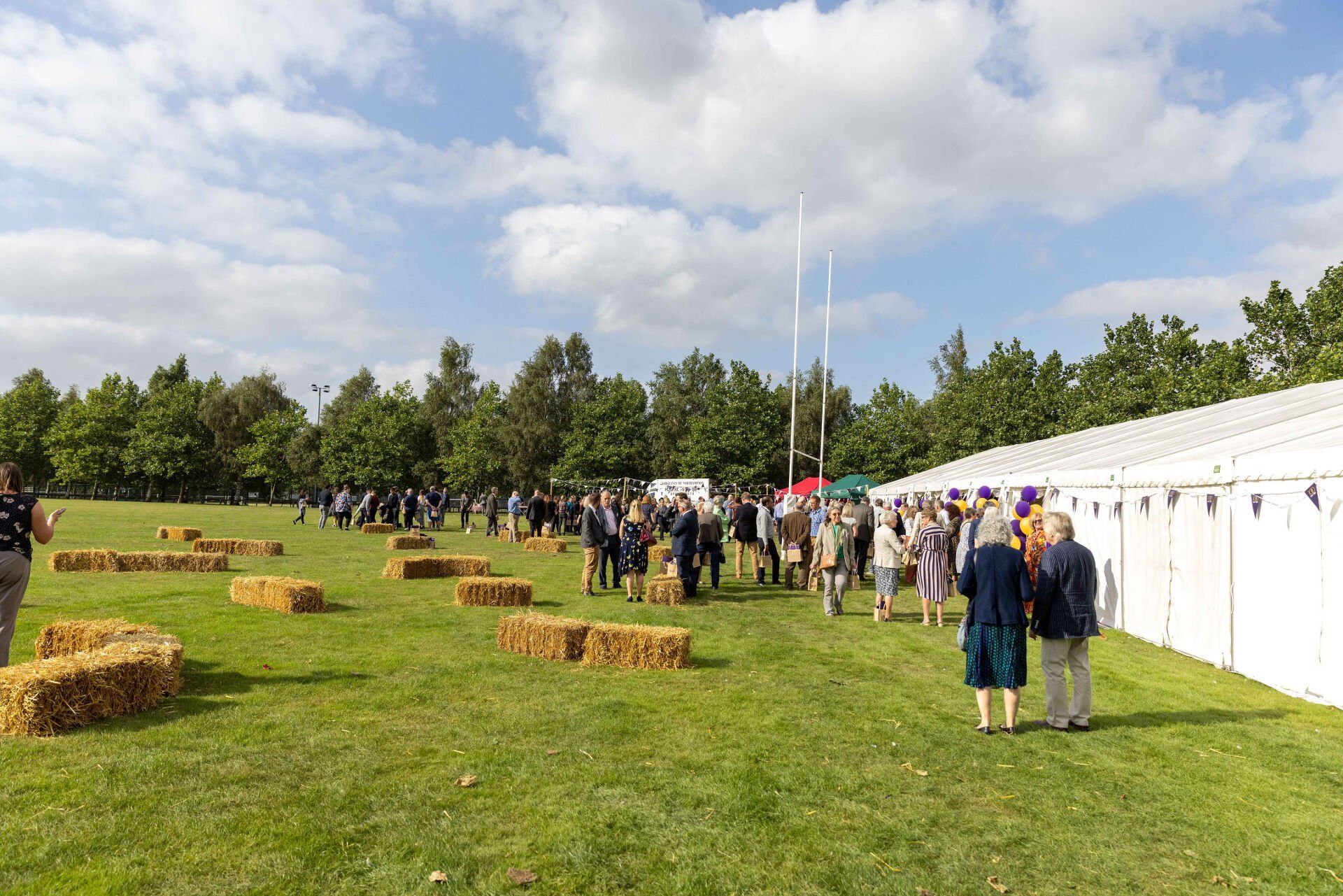 People gathered on a grassy field with hay bales and a large white tent under a cloudy sky.
