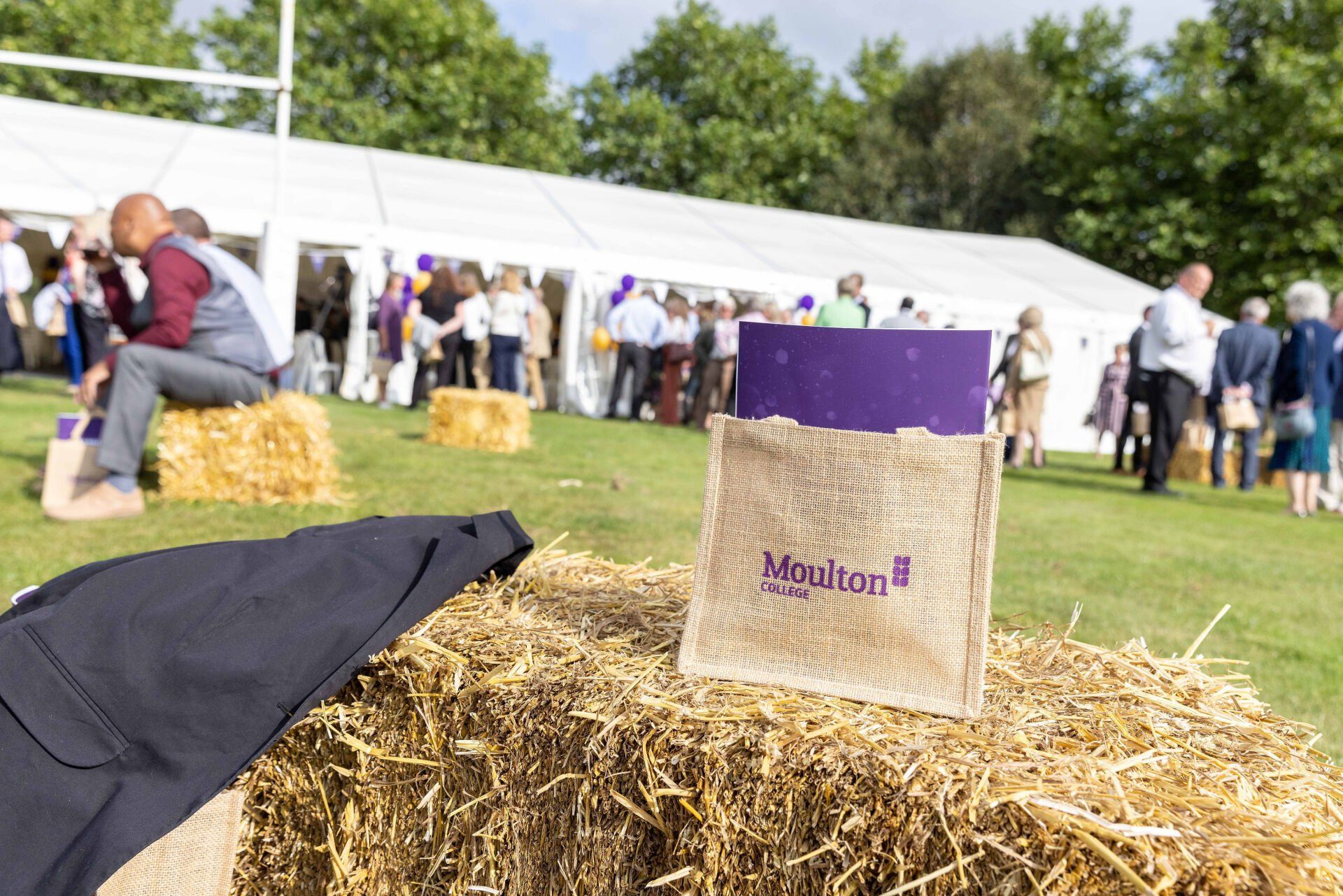 Hay bale with a purple branded item, a crowd near a white tent in a park.
