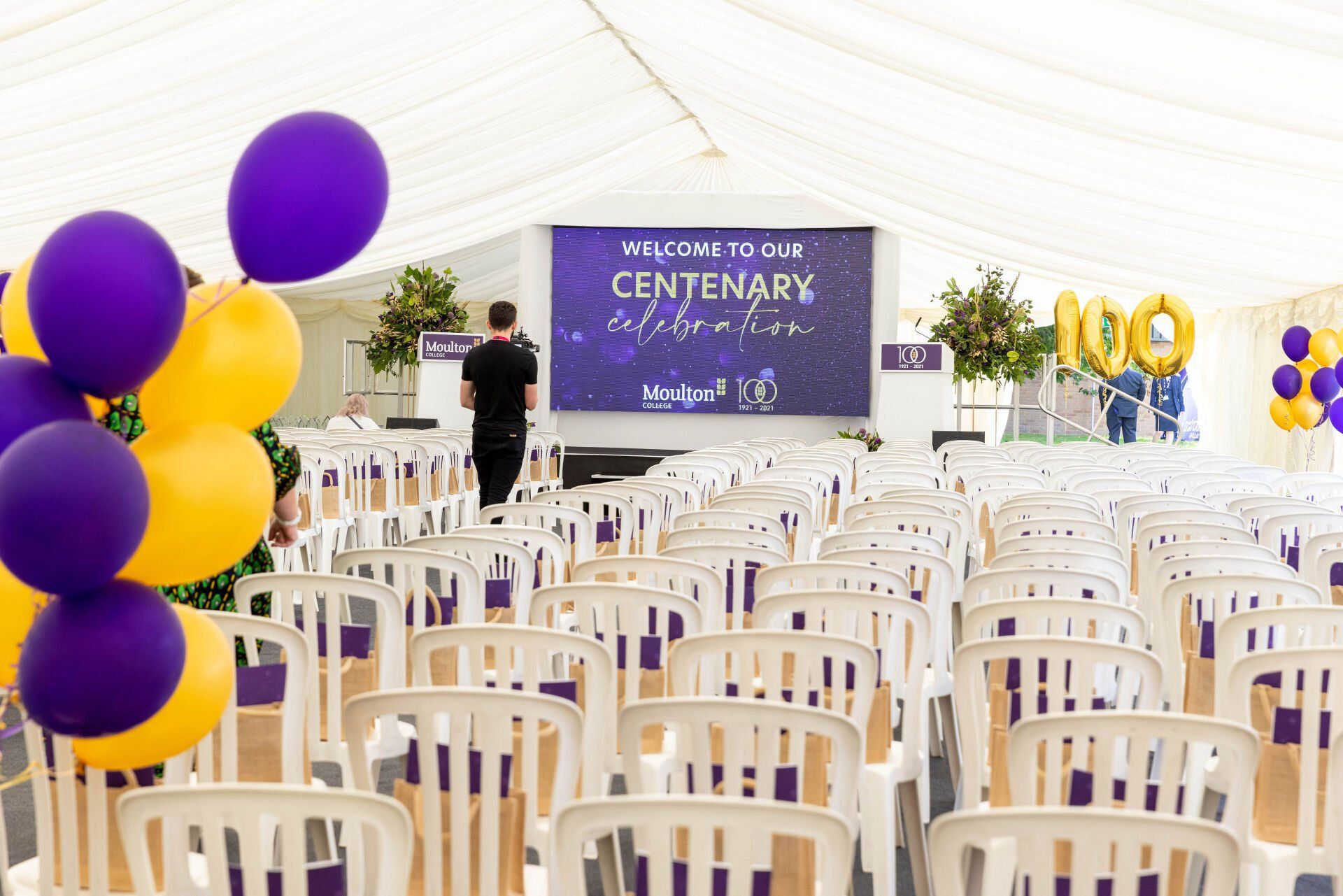 Marquees for Graduation Events White chairs face a stage in a tent, decorated for a centenary. Balloons are purple and yellow.