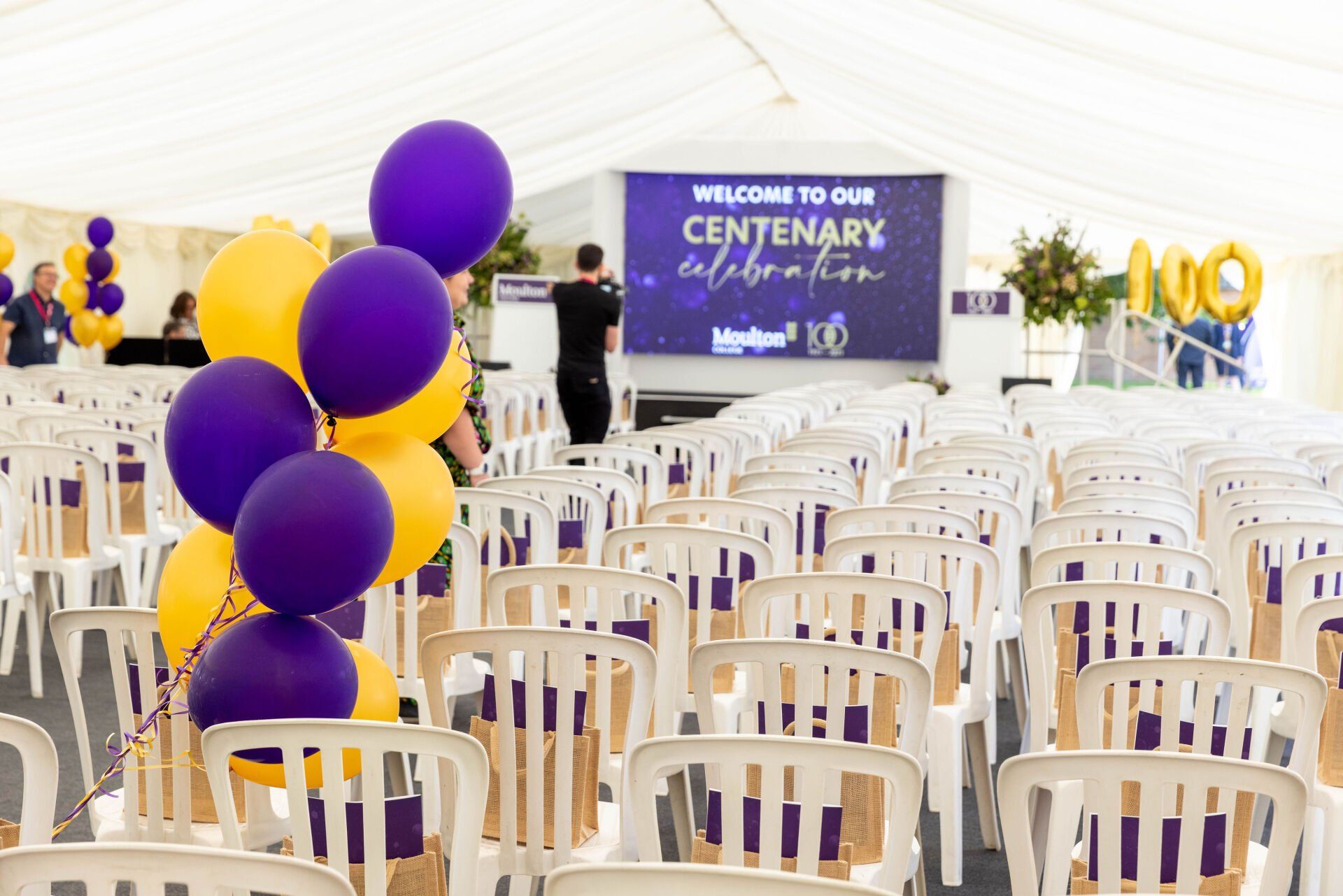 Purple and yellow balloons in front of an event tent with rows of chairs, a screen, and a