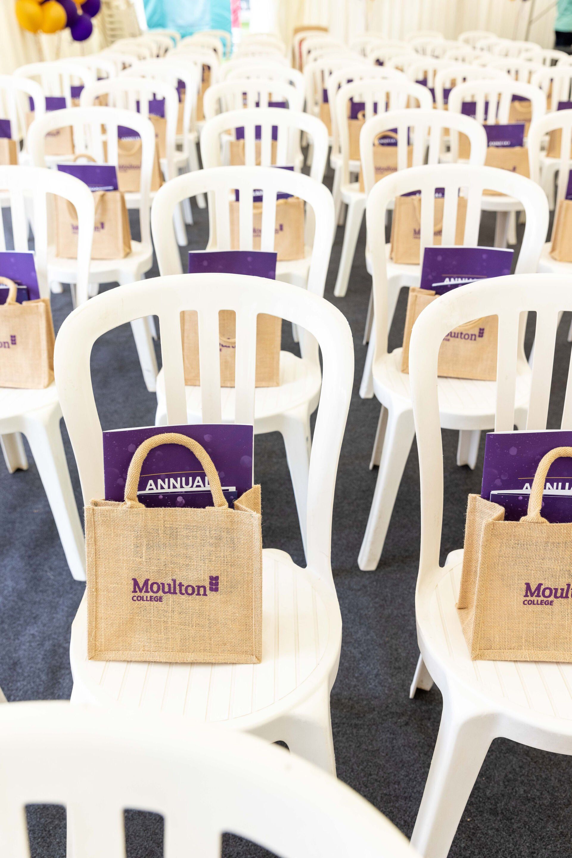 Rows of white chairs with tote bags and purple notebooks; event setting.