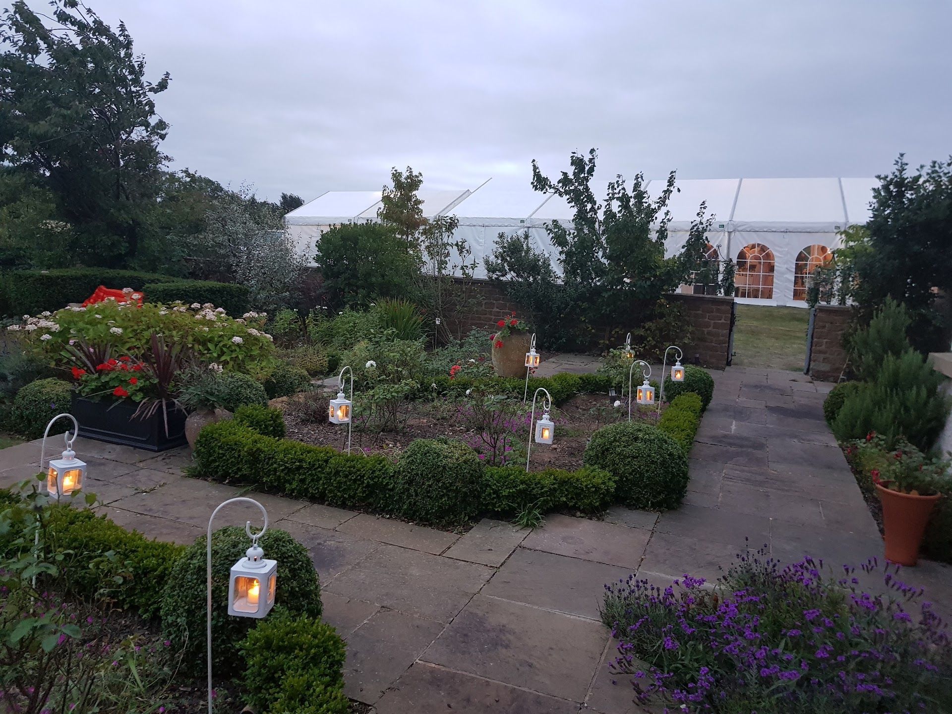 Formal garden with lit lanterns, hedge rows, and a white tent in the background under an overcast sky.