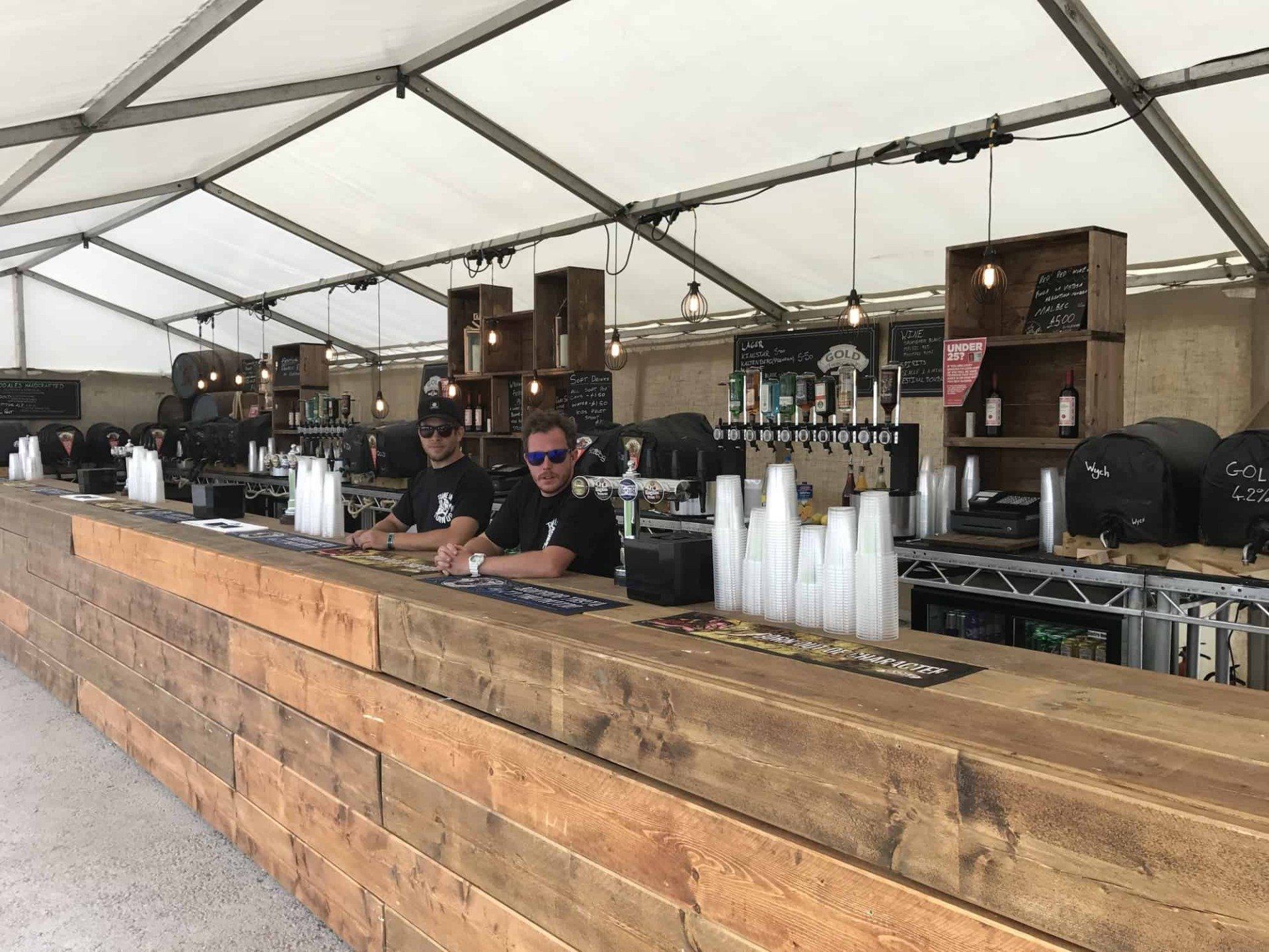 Two people behind a wooden bar inside a tent, serving drinks.