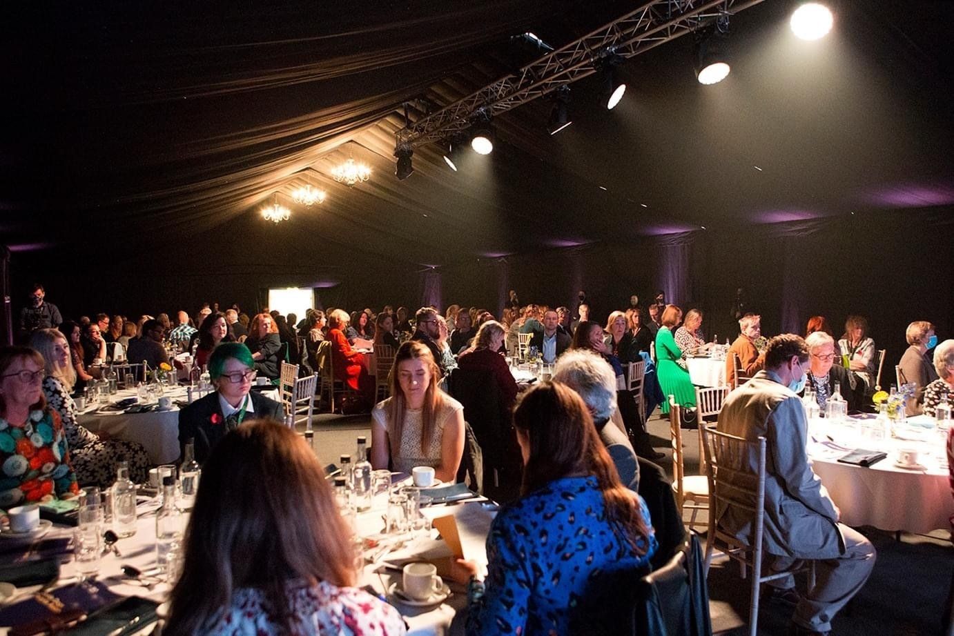 Large event: people seated at round tables.  A stage is lit in front; black ceiling, lights.
